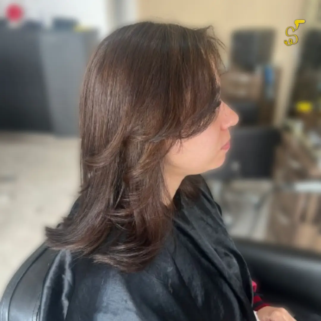 Mulher de perfil com corte de cabelo médio em camadas e franja lateral, na cor castanho escuro, finalizado com escova modelada em salão de beleza.