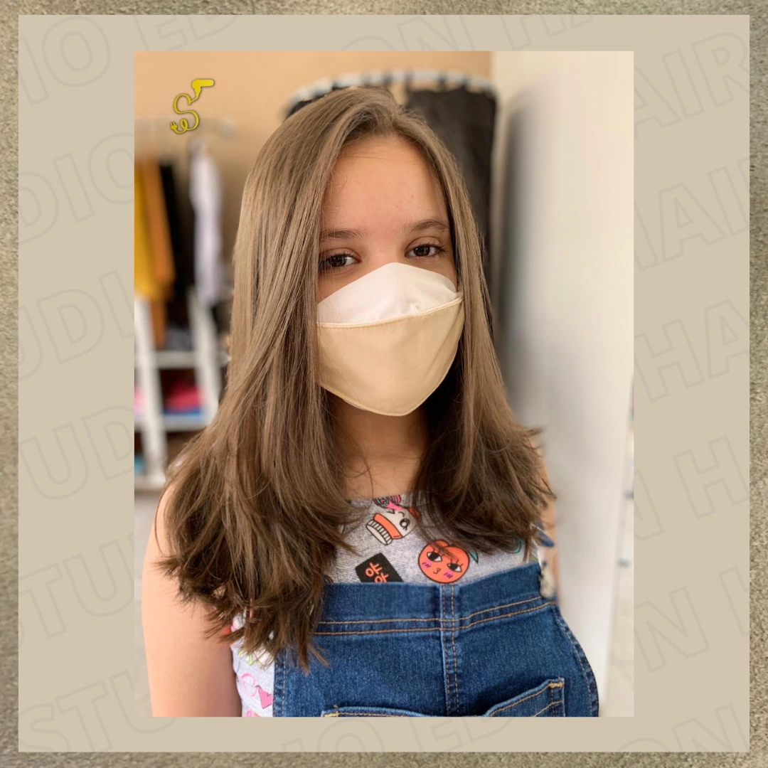Menina usando máscara de proteção exibindo corte de cabelo médio liso, na cor castanho claro natural, com pontas levemente repicadas e franja lateral longa.